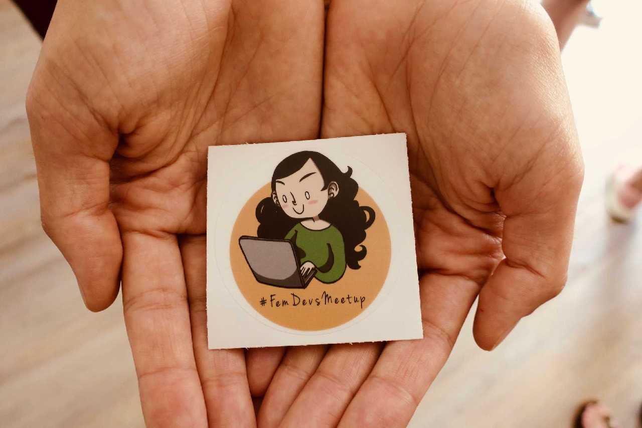 Das Logo des #FemDevsMeetups auf einem Sticker präsentiert in zwei Händen.